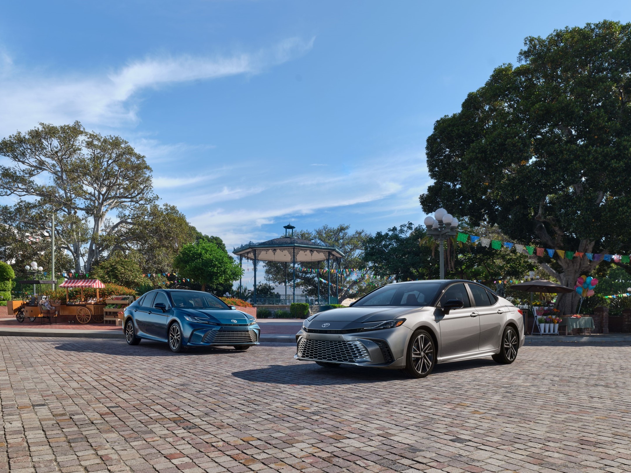 2025 Toyota Camry XLE in Reservoir Blue next to a 2025 Toyota Camry XSE in Celestial Silver Metallic both with Hammerhead Headlamps on display in an open park